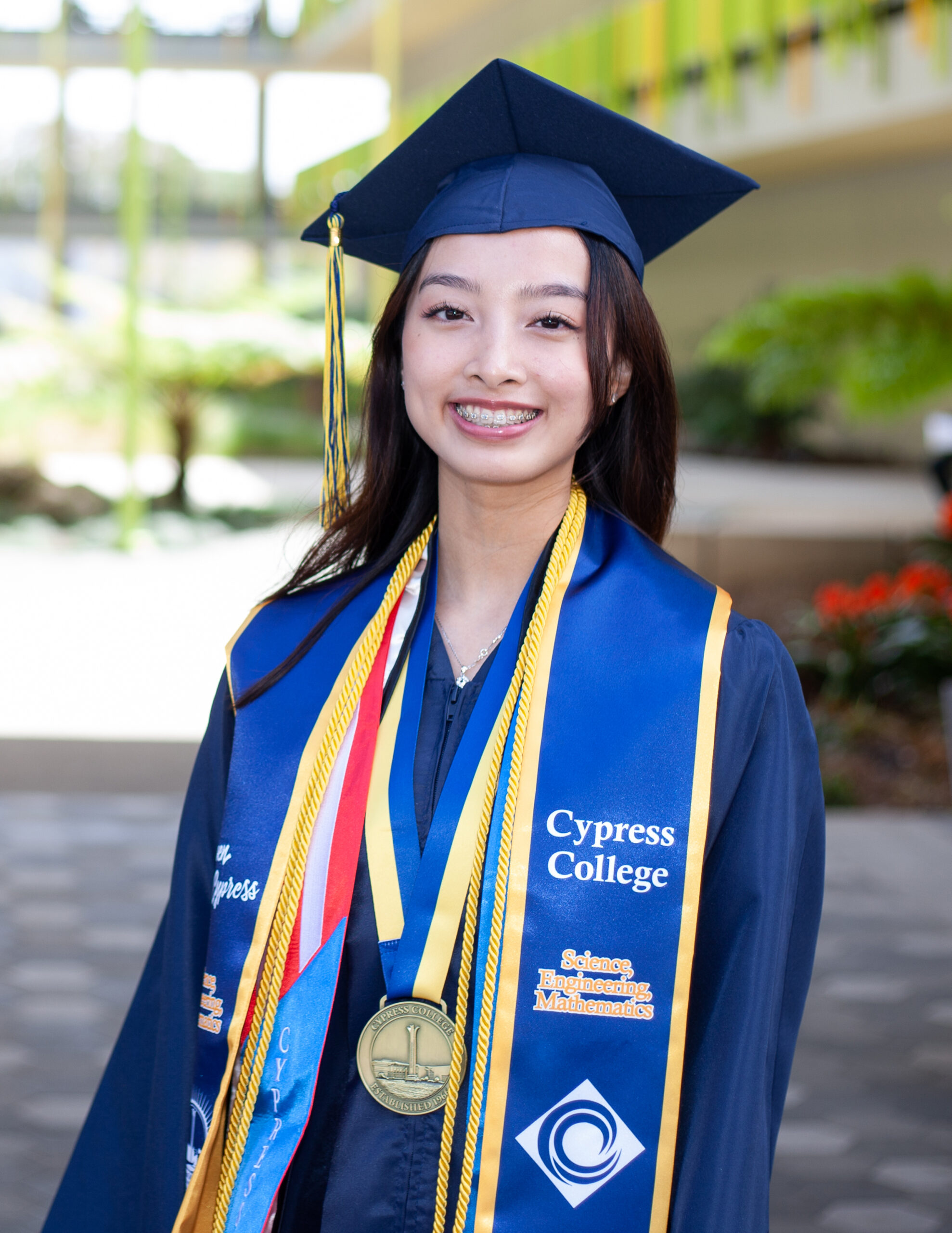Portrait of Mia Nguyen, the 2023 Presidential Scholar of Distinction for the Science, Engineering, Mathematics pathway, wearing regalia with the SEM building in the background.
