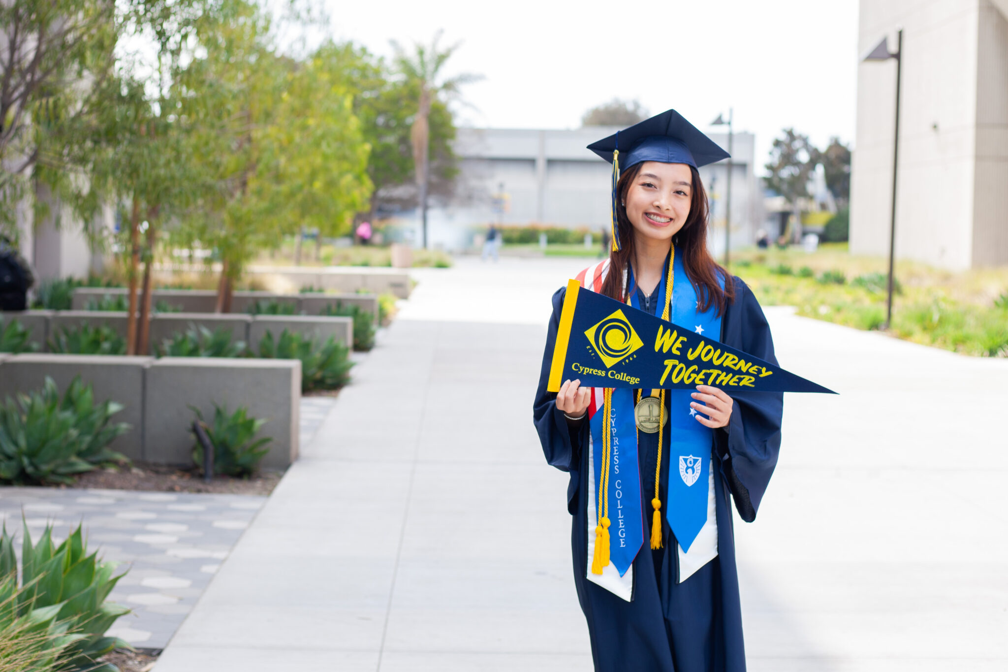 Portrait of Mia Nguyen, the 2023 Presidential Scholar of Distinction for the Science, Engineering, Mathematics pathway, wearing regalia and holding a Cypress College "We Journey Together" pennant with the pond in the background.