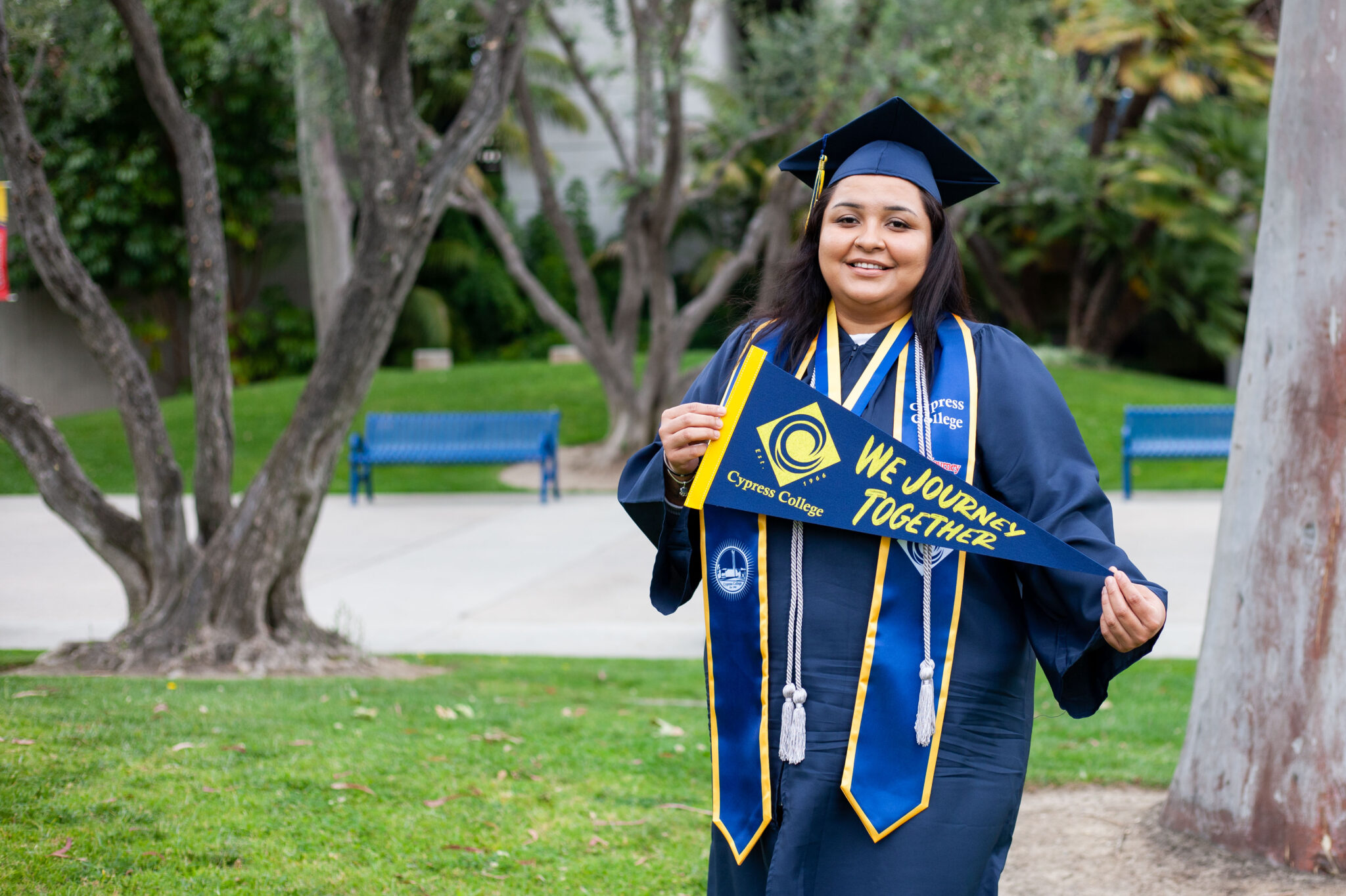 Portrait of Azusena Zamarripa, the 2023 Presidential Scholar of Distinction for the We Journey Together pathway, wearing regalia and holding a Cypress College "We Journey Together" pennant with CCCPLX and greenery in the background.