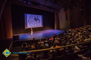 Audience watching woman on stage with cartoon on big screen.