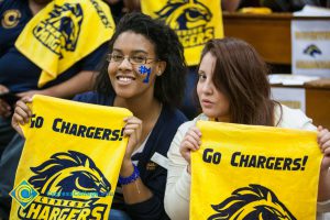 A female fan with glasses and #1 painted on her face in blue, and another young lady hold up yellow Chargers towels.