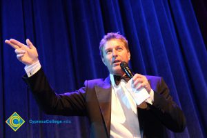 Man in black suit and bowtie holding a microphone and holding hand up.