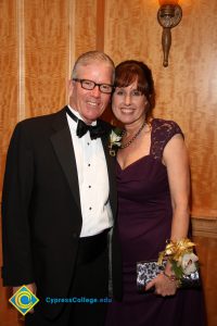 A smiling Bob Simpson in a black tuxedo with his wife Denise who is smiling in a burgundy gown with a wrist corsage.