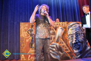 Artist who painted mural of John Wooden speaks at the 39th Annual Americana Awards as a man in a black suit and tie looks on.