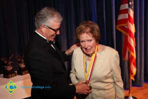A woman in a cream colored outfit with President Bob Simpson as he looks at the award around her neck.