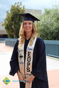 A young lady with blonde hair wearing her cap and gown with an EOPS stole.
