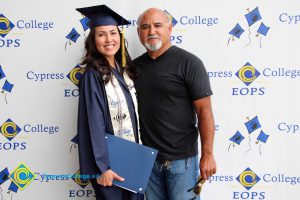 A young lady in graduation cap and gown with a man with grey moustache and beard in a black shirt and blue jeans.