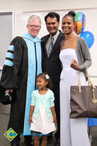 Presidentt Bob Simpson, Peter Mathews and a woman with hair pulled back in a bun wearing a grey sweater and white jumpsuit and a little girl with a dress and flower in her hair.