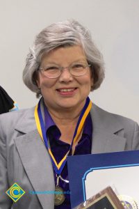 Woman with glasses, grey hair and grey jacket and blue blouse, smiling.
