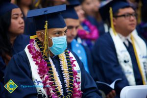 A student in cap and gown with several leis, wearing a surgical mask during the 48th Commencement.