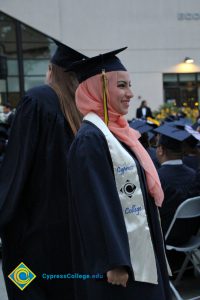 A woman in graduation cap and gown and a coral hijab smiling at during commencement.