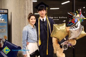 A young woman in a blue denim blouse and white skirt with a young man in a graduation cap and gown holding balloons and a large stuffed animal.