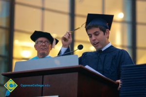 A young man in cap and gown holding his tassel while speaking at the 48th Commencement.