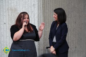 A young woman in a black and white polka dot dress with black sweater holding a microphone while another woman with short brown hair nad black suit looks on with a smile.