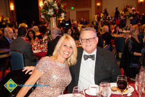 Blond woman in a tan dress at the dining table with a man in a suit and tie wearing glasses.