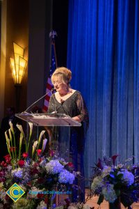 Woman wearing a black evening dress speaking on stage at the podium.