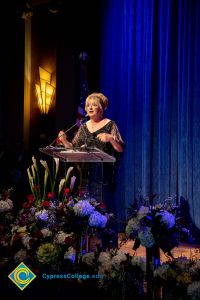 Woman wearing a black evening dress speaking on stage at the podium.