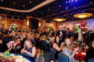 Audience seated at dinner tables applauding.