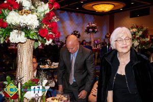 Bald gentleman wearing suit and tie looking down and woman with grey hair, glasses and black dress with eyes closed.