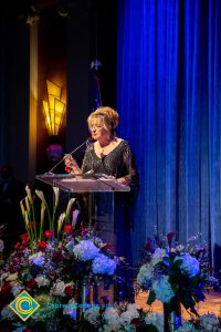 Woman wearing a black evening dress speaking on stage at the podium.