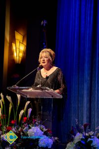 Woman wearing a black evening dress speaking on stage at the podium.