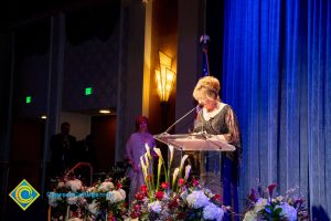 Woman wearing a black evening dress speaking on stage at the podium.