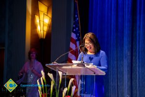 Woman with short dark hair and glasses wearing a blue dress.