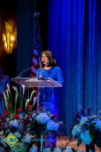 Woman with short dark hair and glasses wearing a blue dress speaking at the podium.