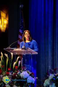 Woman with short dark hair and glasses wearing a blue dress speaking at the podium.