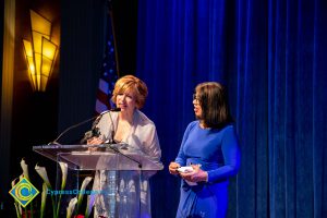 President, JoAnna Schilling speaking at podium with a woman with a blue dress looking on.