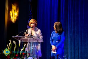 President, JoAnna Schilling speaking at podium with a woman with a blue dress looking on.