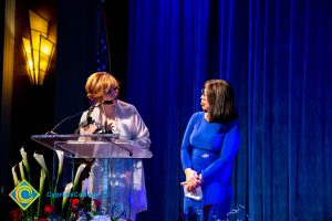 President, JoAnna Schilling speaking at podium with a woman with a blue dress looking on.