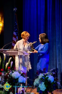 President, JoAnna Schilling speaking at podium with a woman with a blue dress looking on.