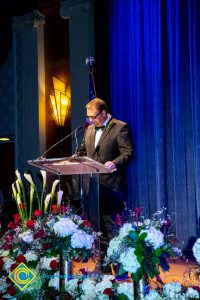 A man in a black suit and tie speaking at the podium.