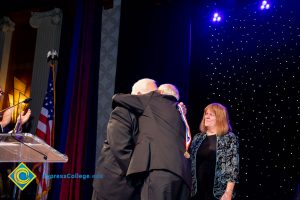 A woman is looking on as two men embrace during the 44th Annual Americana Awards.