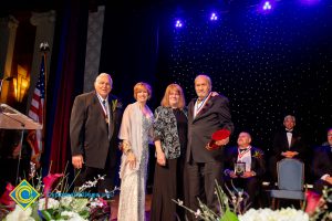 Two men in suits with awards and Dr. JoAnna Schilling and another woman in a black and white floral jacket and black dress.