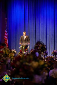 A man in a black suit and tie speaking at the podium.