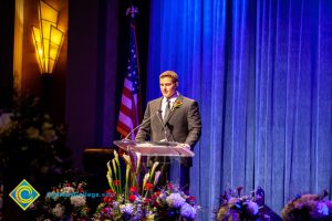 A man in a black suit and tie speaking at the podium.