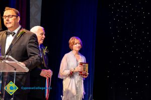 Man standing at the podium with JoAnna Schilling and another man hold awards.