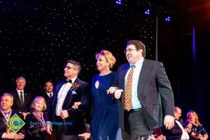 Woman in a blue dress being escorted on stage by two men in suits.