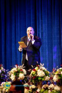 A man in a black suit and tie speaking at the podium.