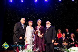 JoAnna Schilling on stage with a woman in a burgundy dress holding an award and two men in suits.