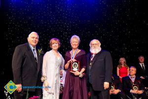 JoAnna Schilling on stage with a woman in a burgundy dress holding an award and two men in suits.