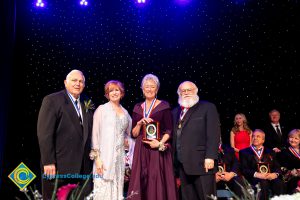 JoAnna Schilling on stage with a woman in a burgundy dress holding an award and two men in suits.