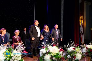 Two men in suits escort a woman in a black dress to podium.