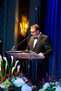 A man in a black suit and tie speaking at the podium.