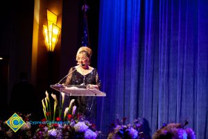 Woman wearing a black evening dress speaking on stage at the podium.