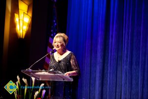 Woman wearing a black evening dress speaking on stage at the podium.