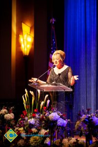 Woman wearing a black evening dress speaking on stage at the podium.