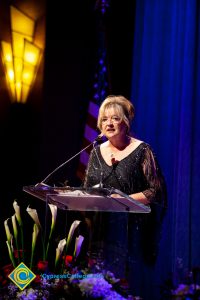 Woman wearing a black evening dress speaking on stage at the podium.